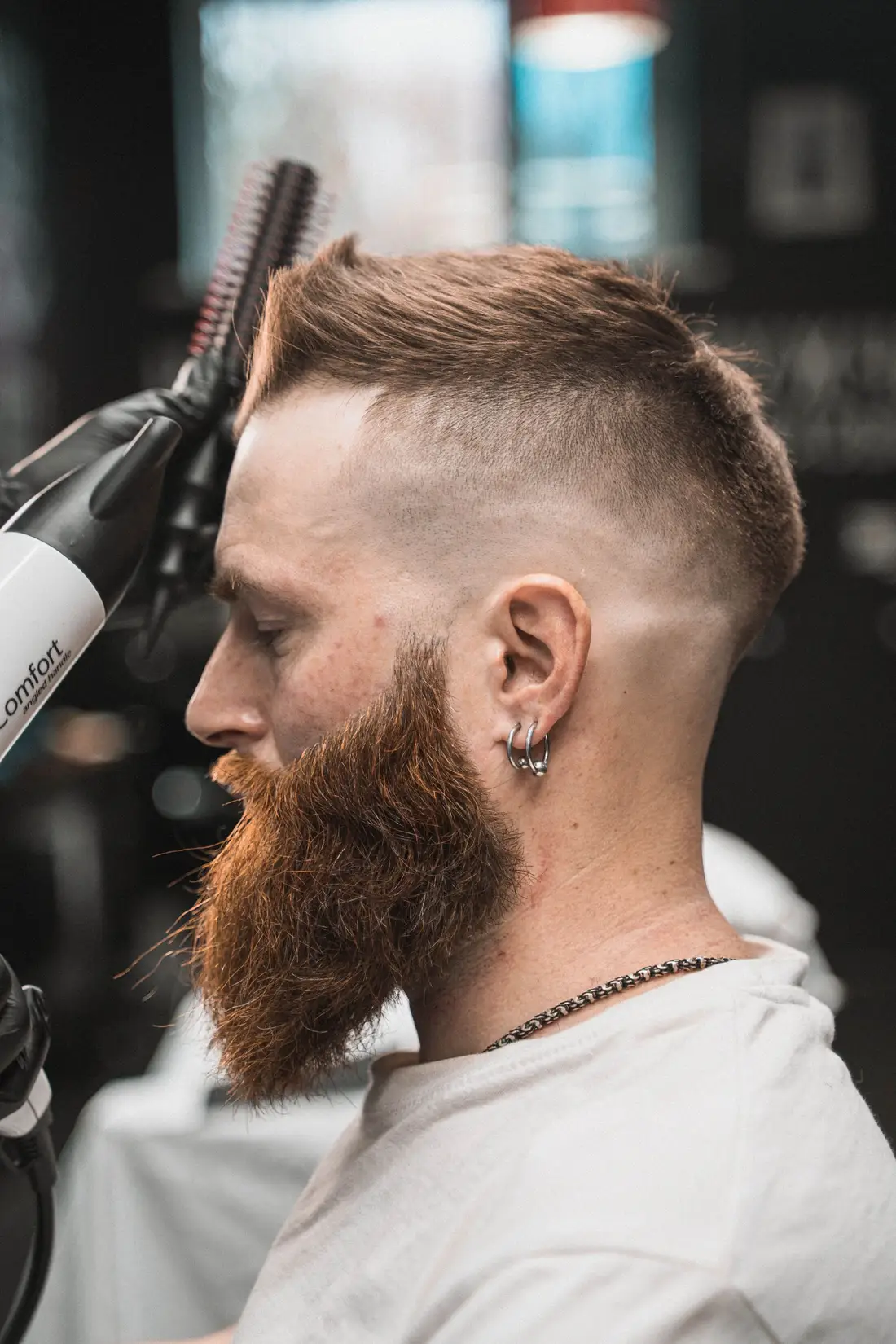 Profil mężczyzny z rudą brodą po strzyżeniu z fade na boku głowy, barber suszy włosy suszarką.