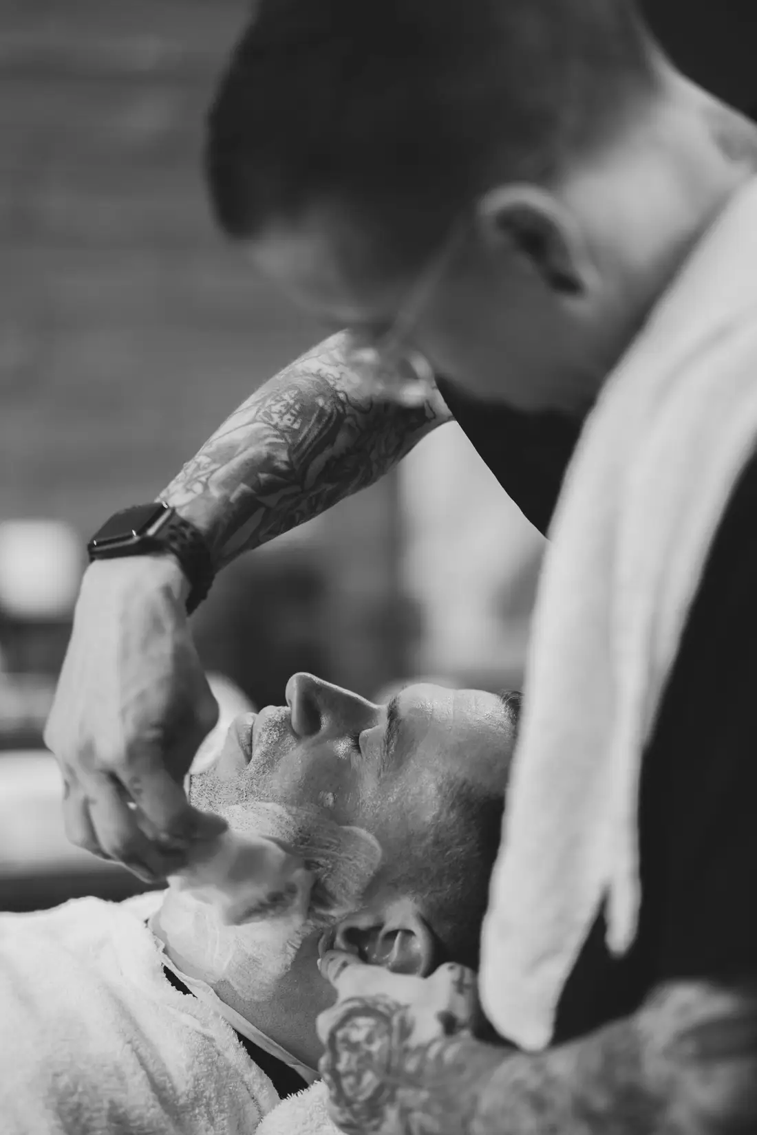 Czarno-białe ujęcie: barber goli brzytwą leżącego klienta z ręcznikiem na czole.