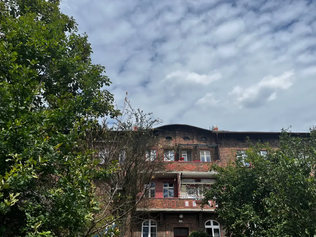 Ceglany budynek mieszkalny w Nikiszowcu widziany zza drzew, z rzędem okien w czerwonych obramowaniach.