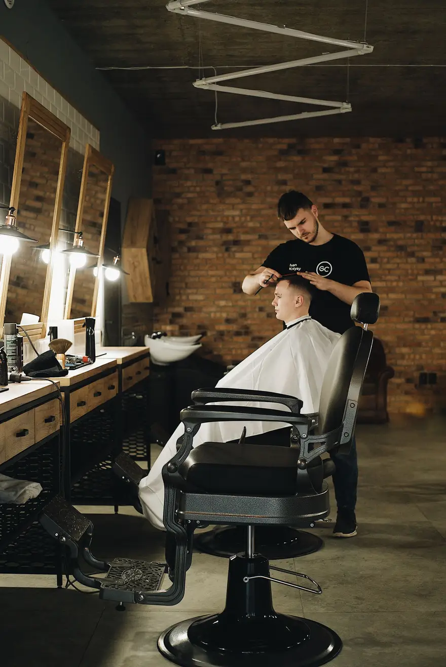 Wnętrze barber shopu wieczorem: ceglana ściana, lustra w ramach i barber przy pracy nad klientem w fotelu.