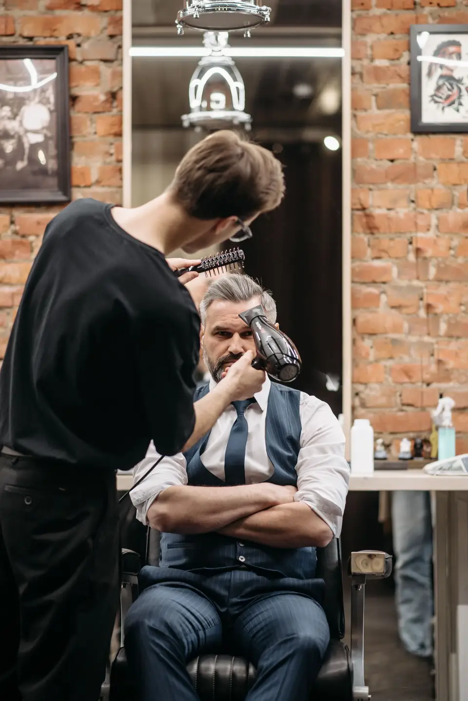 Barber suszy i układa szczotką włosy klienta siedzącego w fotelu przy ceglanej ścianie.