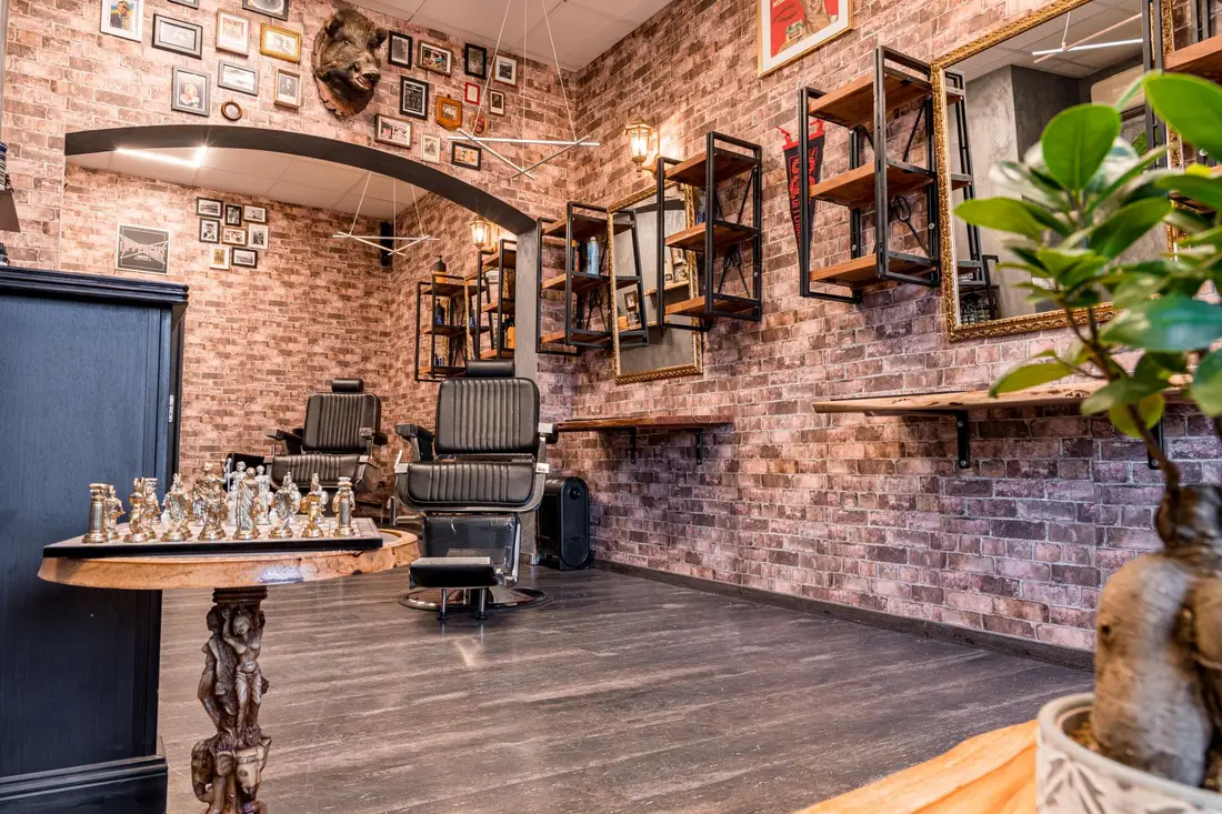 Dwa fotele barberskie ustawione przy ceglanej ścianie, obok drewniane półki z narzędziami.