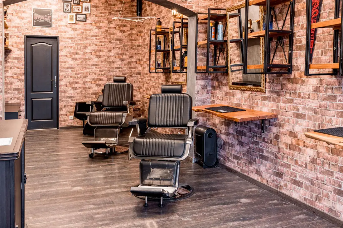 Wnętrze barber shopu: dwa fotele, drewniana podłoga i ceglana ściana z lustrami oraz ramkami.